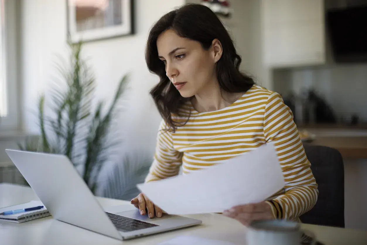 Woman in a striped yellow-and-white top reviewing a printed document while working on a laptop at a home desk.