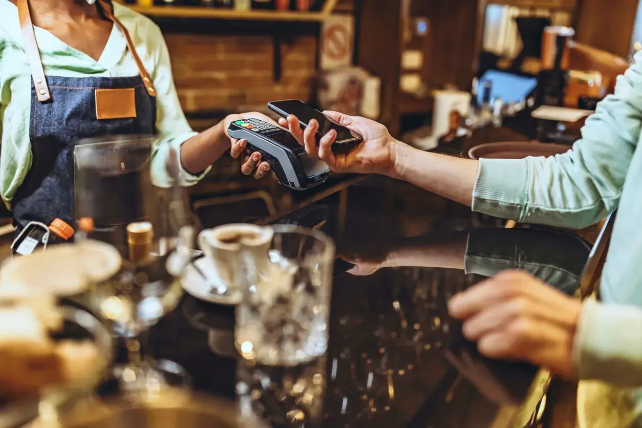 A customer pays contactlessly with a smartphone at a café counter while a barista holds the card terminal.